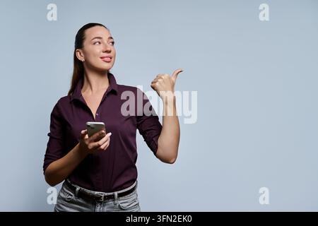 Giovane donna che tiene uno smartphone e indica con il pollice di lato sorridendo Foto Stock
