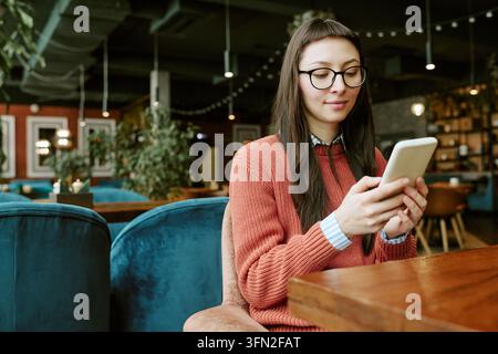 Donna caucasica con occhiali che utilizza uno smartphone seduta in un caffè Foto Stock