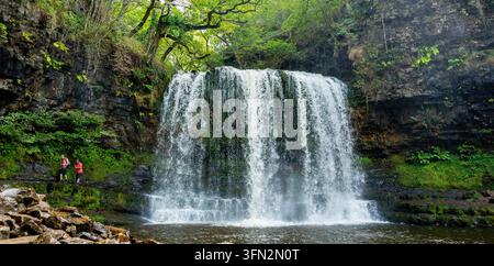Una vista panoramica di una cascata che scende su scogliere di pietra scura con due persone che percorrono un sentiero stretto circondato da una vivace foresta Foto Stock