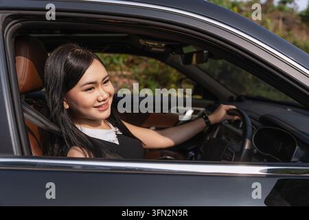 Giovane donna asiatica sorridente seduta sul sedile del conducente che tiene il volante. Foto Stock