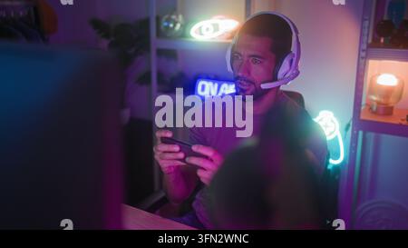 Uomo con auricolare e microfono tiene uno smartphone con entrambe le mani, gesticola con la mano sinistra e sorride seduto in uno studio di gaming al neon; eccitazione Foto Stock