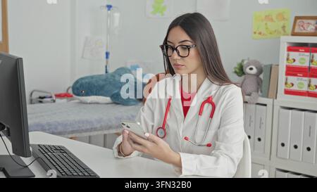Giovane medico con stetoscopio che tiene lo smartphone, le mani visibili e scorrono mentre si siede alla scrivania del computer in un edificio; concentrazione tranquilla. Foto Stock