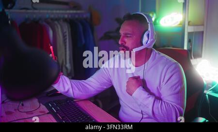 Uomo con auricolari tiene uno smartphone e gesticola con la mano in un setup da gioco in studio; gioia del gioco online. Foto Stock
