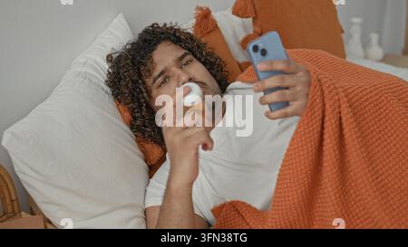 Giovane indiano giacente a letto sotto coperta arancione tiene smartphone, tocca la gola con mano e i principi accanto alla bottiglia di pillola in studio; preoccupazione per la salute. Foto Stock