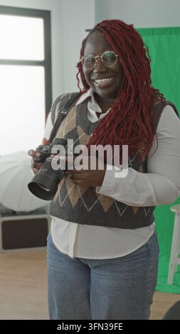 La donna tiene in mano la fotocamera e sorride mentre regola l'obiettivo in uno studio fotografico luminoso, indossa occhiali rotondi e un gilet argyle; creativo j Foto Stock