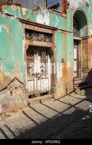 Facciata turchese consumata ad Ayvalık, Turchia, con una porta di legno chiusa a chiave, griglie di ferro, intonaco scrostato, ingresso ad arco e ombre intense. Foto Stock