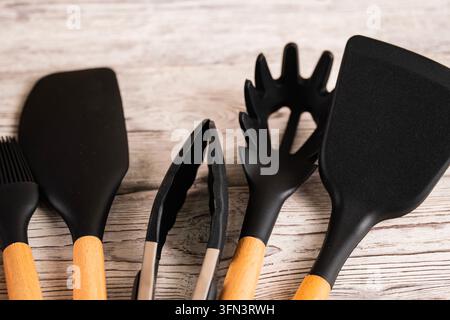 Set di utensili da cucina in silicone nero con manici in legno disposti su uno sfondo di legno chiaro. Strumenti culinari moderni per cucinare e infornare a casa Foto Stock