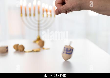 Un primo piano cattura una mano che fa girare un dreidel di legno in un ambientazione di Chanukkah, la luce delle candele proietta un caldo bagliore sulla venatura del legno, con una menorah sfocata nel t Foto Stock