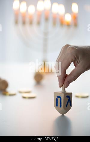Primo piano di una mano che tiene un dreidel di legno su un tavolo bianco, mostrando lettere ebraiche mentre il giocoliere poggia in posizione verticale, con una luce naturale soffusa Foto Stock