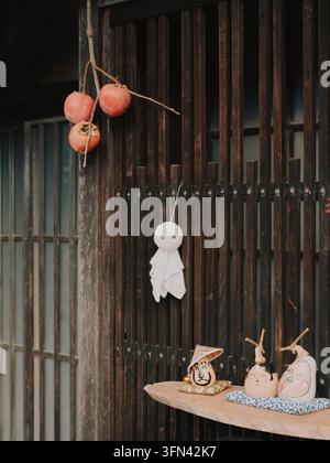 Persimoni essiccati (hoshigaki) appesi accanto a una bambola Teru Teru Bozu e a figurine di gatti portafortuna fuori da una casa tradizionale in legno a Nagano, Giappone. Foto Stock