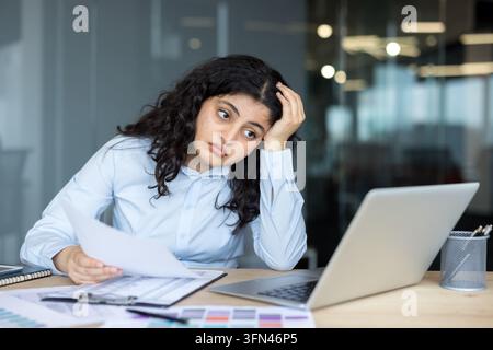 Giovane donna d'affari con capelli ricci scuri seduta alla scrivania dell'ufficio, tiene documenti e sembra stanca e sopraffatta mentre vive stress lavorativo e burnout Foto Stock