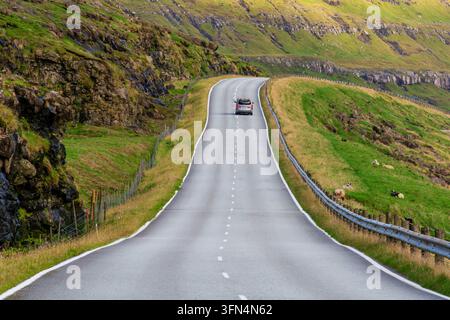 Vista di un'auto che percorre una strada tortuosa attraverso le colline verdi e rocciose delle Isole Fær Øer. Foto Stock