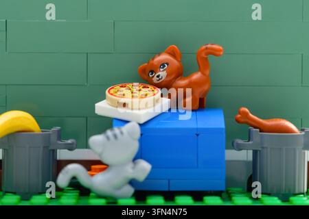 Tambov, Federazione Russa - 03 agosto 2026 Gatto di Lego color zenzero in piedi su un cassonetto blu accanto a una pizza, mentre un gatto grigio e bidoni della spazzatura con cibo lo Foto Stock