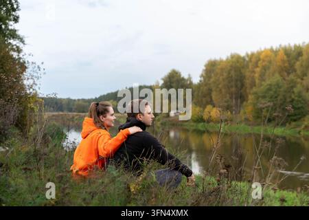 Giovane uomo e donna seduti insieme su una riva erbosa del fiume che godono di un tranquillo paesaggio autunnale, del fogliame della foresta e dell'acqua calma Foto Stock