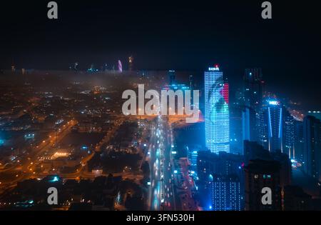 Kuwait City, Kuwait - 10 dicembre 2023: Veduta aerea della nebbia sulla città di notte, con tracce di luce dei semafori su una strada principale e torri illuminati di blu. Foto Stock