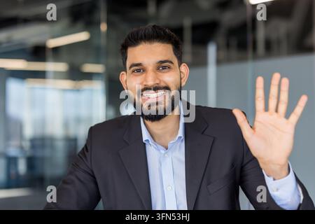 Giovane uomo d'affari indiano che sorride e agita la mano, salutando durante una videoconferenza o un incontro virtuale da un ambiente moderno di ufficio aziendale, trasmettendo cordialità e connessione Foto Stock