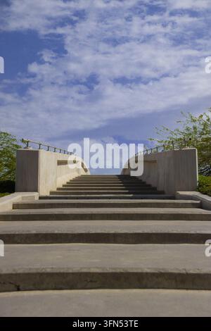 Vista ad angolazione bassa verso ampi gradini di cemento che portano a un ponte pedonale curvo con ringhiere metalliche sotto un cielo azzurro con nuvole bianche, Kuwait Foto Stock