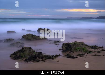 Il mare calmo circonda gli scogli su una spiaggia sabbiosa del Pembrokeshire sotto un cielo serale nuvoloso con luce calda lungo l'orizzonte. Foto Stock