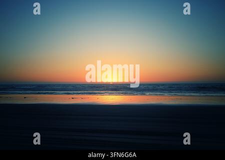 Tramonto sull'oceano dorato con cielo a gradiente di colori vivaci e riflessi sulla sabbia bagnata della spiaggia Foto Stock