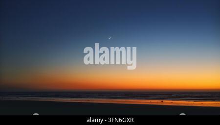 Luna crescente che brilla in un cielo serale blu profondo sopra un tramonto sull'oceano dorato e le onde Foto Stock