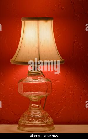 Una lampada da tavolo elettrica antica illuminata con base in vetro trasparente vicino a un muro rosso Foto Stock