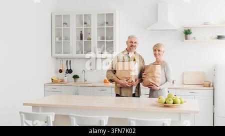 Coppia sposata felice che torna a casa dopo la spesa e svuota insieme borse di carta con la spesa in cucina, famiglia e relazione. Anziano sorridente Foto Stock