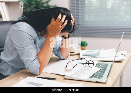 Donna d'affari sopraffatta che tiene la testa a una scrivania con laptop, calcolatrice e rapporti, mostrando burnout, stress lavorativo, pressione delle scadenze, w finanziaria Foto Stock