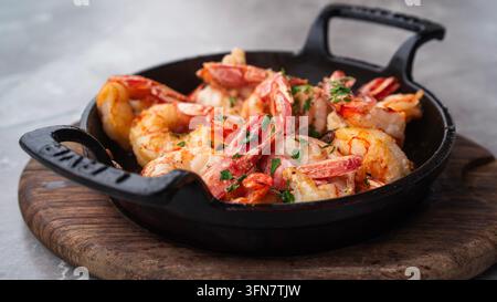 Deliziose gamberetti sfrigolanti saltati con aglio, burro e prezzemolo fresco, serviti caldi in una padella di ghisa nera su un vassoio di legno rustico. Perfetto Foto Stock