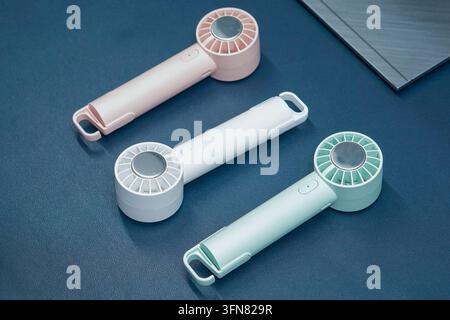 Tre ventole elettriche portatili moderne a mano in colori pastello disposte su una superficie da scrivania blu scuro. Foto Stock