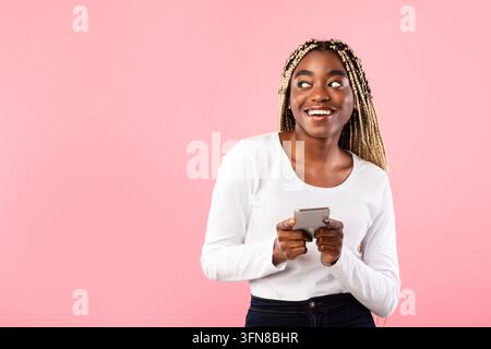 Espressione gioiosa. Adolescente nera sorridente che tiene uno smartphone mentre interagisce con il dispositivo in un ambiente studio luminoso. Concetto di tecnologia moderna Foto Stock