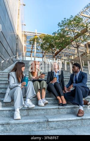Gruppo diversificato di professionisti d'affari seduti sulle scale moderne della città, godendosi una pausa caffè, conversando e collaborando durante un momento rilassato ou… Foto Stock