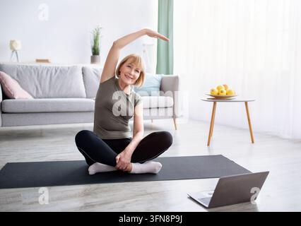 Esercizi di flessibilità per una vita più sana. Donna anziana atletica che fa yoga davanti al laptop durante l'allenamento a casa. Attraente donna matura che pratica Foto Stock