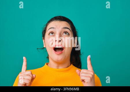 Giovane donna sorpresa che indica verso l'alto in maglione giallo su sfondo verde-azzurro, ritratto con espressione entusiasta Foto Stock