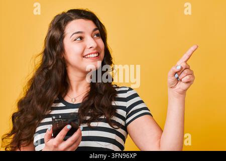 Giovane donna sorridente che tiene uno smartphone puntato verso l'alto su uno sfondo giallo brillante Foto Stock