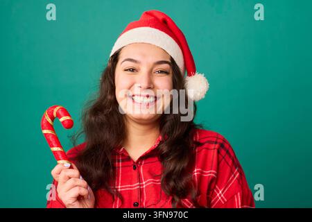 Giovane donna felice con cappello di Babbo Natale che tiene una bastoncino di zucchero e sorride su sfondo verde Foto Stock