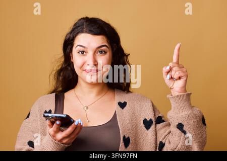 Donna sorridente che tiene uno smartphone puntato verso l'alto con un gesto di idea in un ritratto studio casual Foto Stock