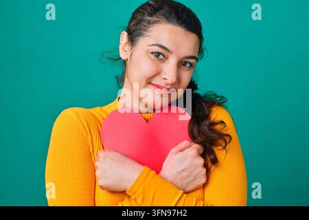 Giovane donna sorridente che abbraccia una carta a forma di cuore rosso in carta, ritratto in primo piano che mostra amore e cura di sé Foto Stock