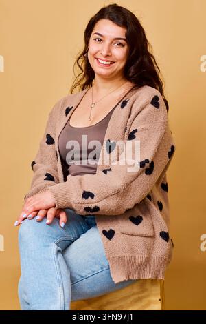 Giovane donna sorridente con cardigan a forma di cuore seduta in jeans per un ritratto in studio casual e accogliente Foto Stock