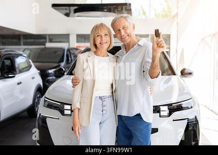 Coppia di anziani felici che si abbracciano accanto a un'auto nuova presso il concessionario, l'uomo tiene la chiave Foto Stock