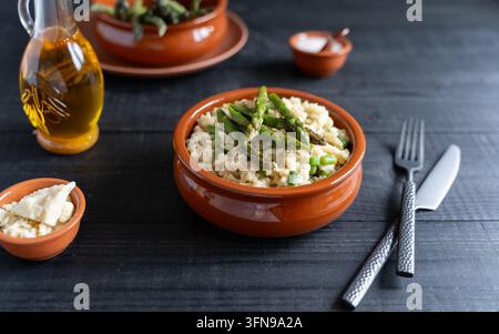 Riso cremoso agli asparagi sormontato di asparagi verdi servito in una ciotola di terracotta con posate e piccoli piatti su un tavolo di legno scuro Foto Stock
