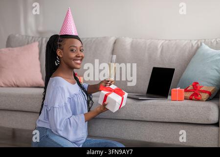 Festeggiamo il compleanno in autoisolamento. Bella giovane donna nera con un bicchiere di champagne e regali che usa un laptop con schermo vuoto, indossa abiti da festa Foto Stock