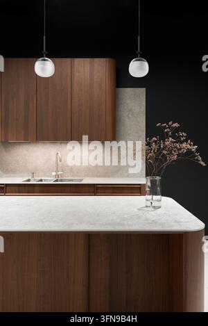 Design elegante della cucina che mostra mobili in legno caldo montati su uno sfondo color carbone. Due lampade a sospensione in vetro trasparente pendono dal soffitto sopra un piano di lavoro in marmo chiaro Foto Stock