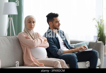 Annoiata donna musulmana incinta offesa a suo marito impegnata con laptop, madre incinta islamica in Hijab sentendosi sola e depressa mentre era seduta su C. Foto Stock
