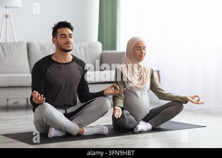 Una coppia musulmana incinta che pratica lo yoga prenatale a casa, meditando insieme Foto Stock