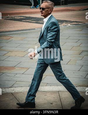 Londra, Regno Unito – 22 giugno 2021: Un uomo dai capelli grigi, in abito blu-grigio, camicia bianca con colletto aperto e occhiali da sole, attraversa un’area lastricata. Indossa un polso Foto Stock