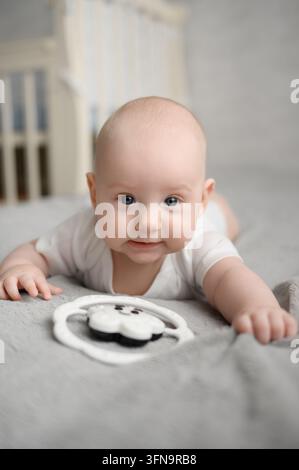 Simpatico neonato maschio che gioca con un giocattolo da masticare durante il tempo sulla pancia. Dolce infante in tutina sdraiato su una coperta morbida nella cameretta. Primi giocattoli, miglio sensoriale Foto Stock
