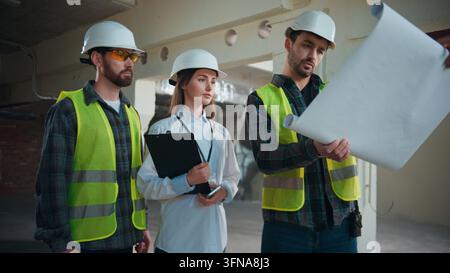 Squadra edile caucasica che pianifica il progetto con la piantina durante la collaborazione, architetta donna che supervisiona operai edili maschi, tre ingegneri Foto Stock