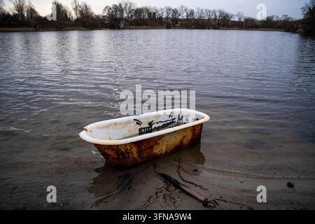 Vista di una vasca da bagno vecchia, arrugginita e coperta di graffiti abbandonata nell'acqua bassa di un lago con alberi ed edifici sullo sfondo a Copenaghen, Danimarca Foto Stock