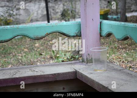 Un primo piano di un bicchiere di plastica monouso su un balcone sporco di un vecchio gazebo in legno colorato al chiuso. Foto Stock