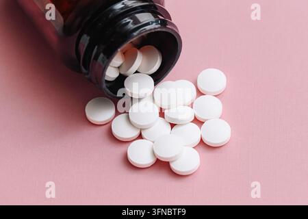 Compresse rotonde bianche che fuoriescono da una bottiglia di medicinali scura su una superficie rosa. Sfondo farmaceutico per farmaci, farmacia, assistenza sanitaria, dosaggio giornaliero. Foto Stock